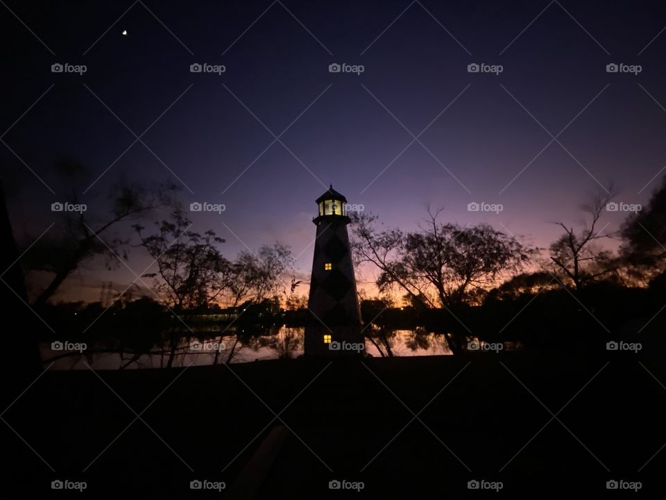 A Beautiful Sun-filled Day Clear Sky’s Overhead this Lighthouse poised for Bright Moonlit Night Filled By Night Colors. Reflections from Peaceful Lake Water with Backlit Twilight hanging on into Night.