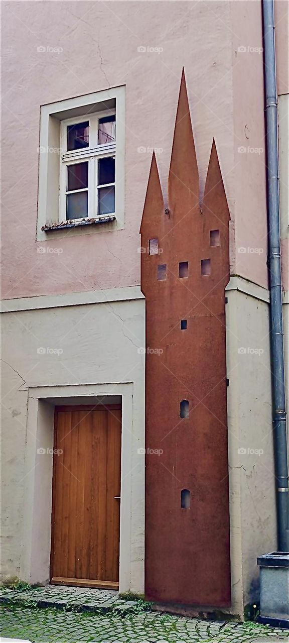 This is a one and a half story high wooden replica of the “Stadtturm”, the city tower of “Straubing” in “Niederbayern” or “Lower Bavaria”, Germany attached to the facade of an old style building in pastel pink. 2024. Hypnotic Productions