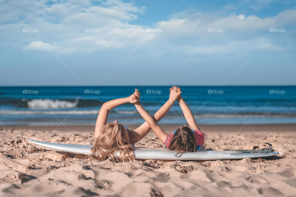 Two girls laying on the surfboard on the beach and holding each other hands 