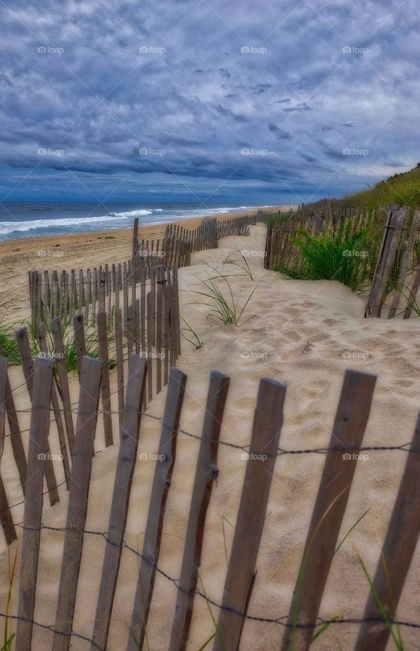 Fenceline leading to the beach, fence along the beach, walking along the seaside, portrait of a seashore, waves rolling onto the beach, cloudy skies on the beach