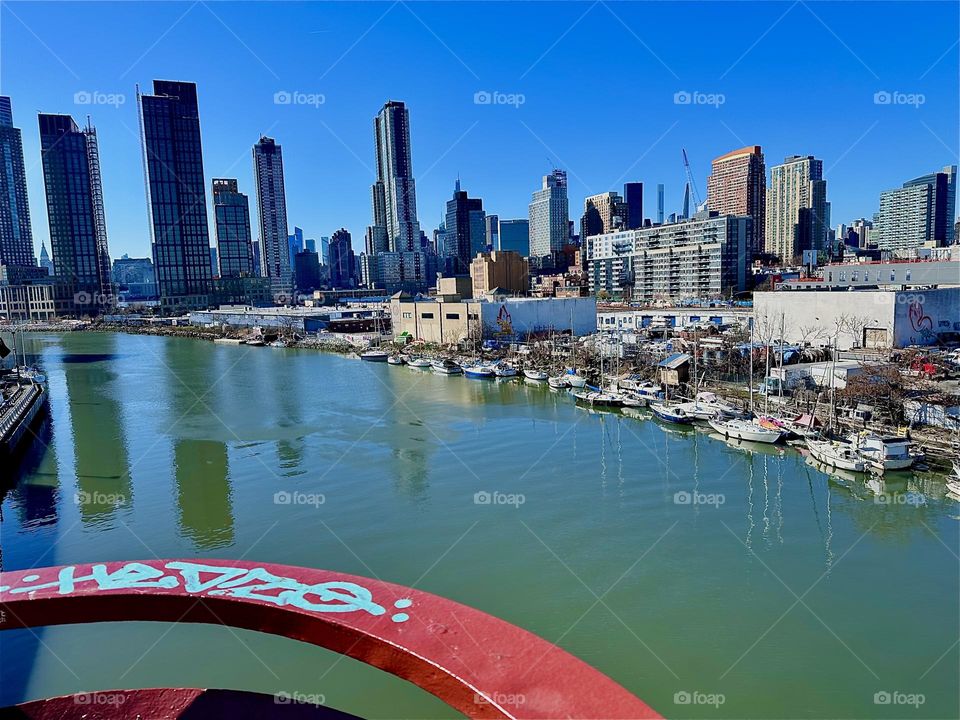 This is beautiful „Newtown Creek“ with all its boats seen from the „Pulaski Bridge“ that connects LIC, Queens to „Greenpoint“, Brooklyn. Across the water of the „East River“ behind LIC we see „Manhattan“. 2024. Hypnotic Productions