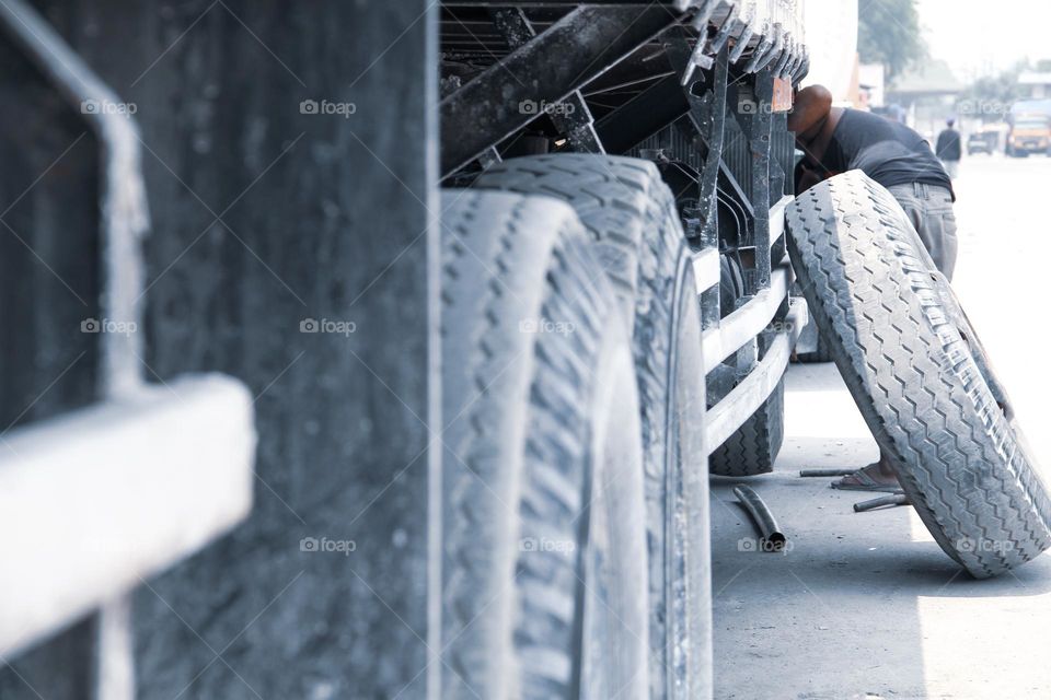 A large truck tire propped against the body of a truck by a man repairing it by the side of the road during the day.