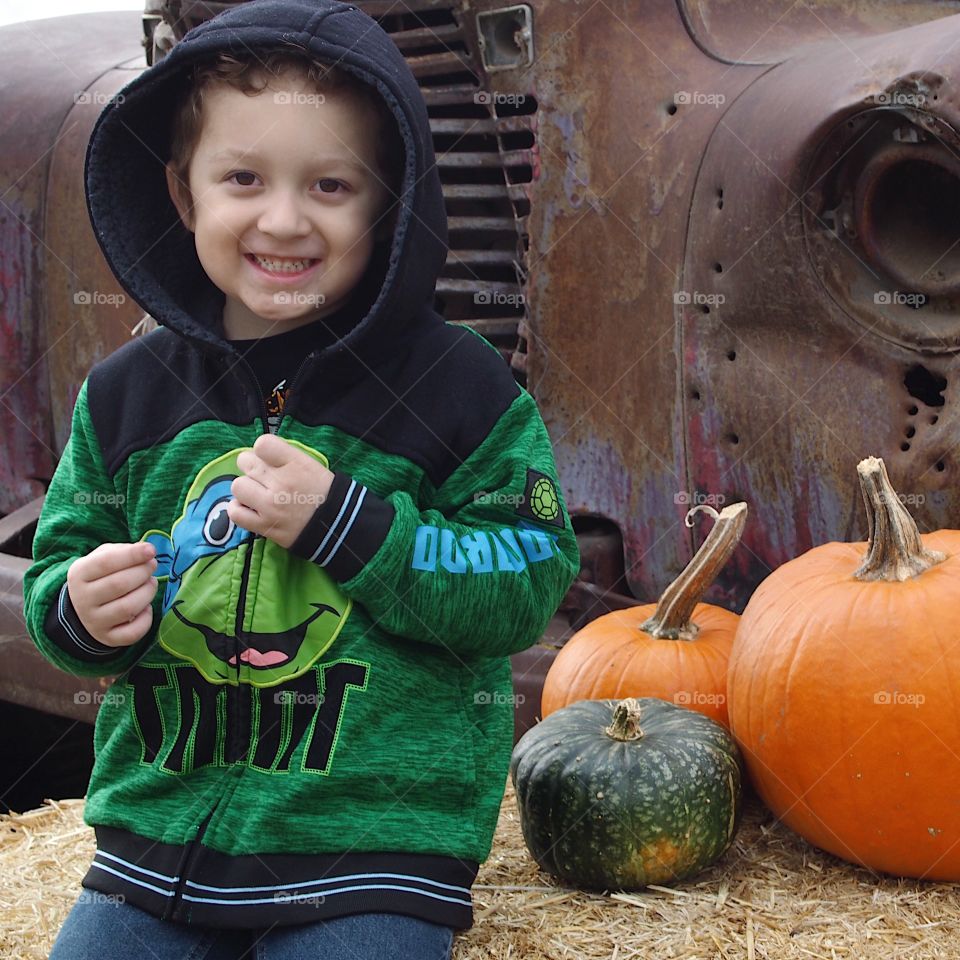 A delighted young boy sitting on a hay bale adorned with pumpkins and an old rusted truck in the background at the Pumpkin Patch in rural Central Oregon on a fall day. 