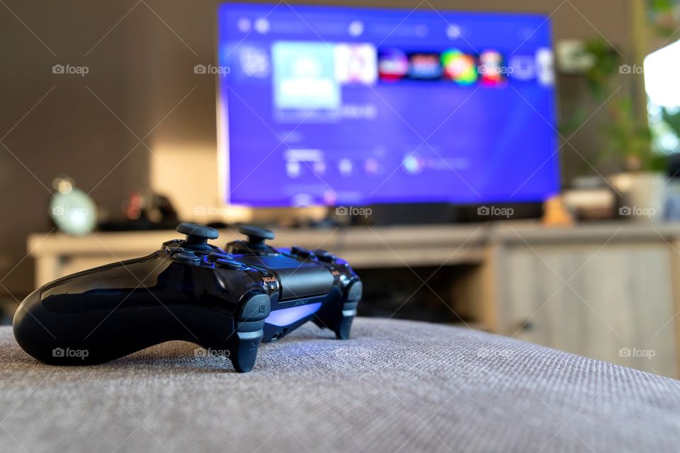 A portrait of a turned on Sony PlayStation 4 controller lying on a couch in front of a television with the PlayStation home screen on it. Playing games is a routine for many people to escape from the stress of the daily life.