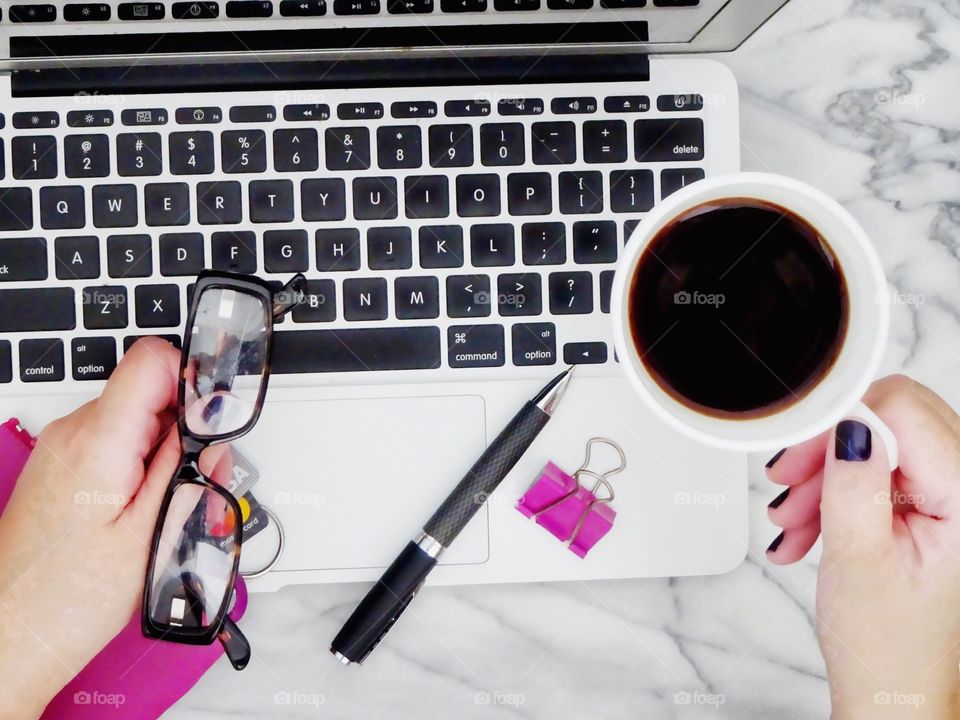 Flat lay overhead shot of laptop, woman's hands holding a cup of coffee and credit cards in pink wallet, glasses, pen and pink paper clip, on a marble table - ready to do some online shopping.