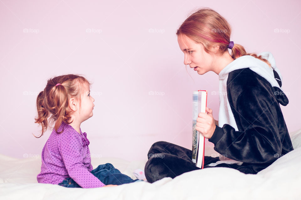 Girl showing the pictures in a book her younger sister while sitting on bed