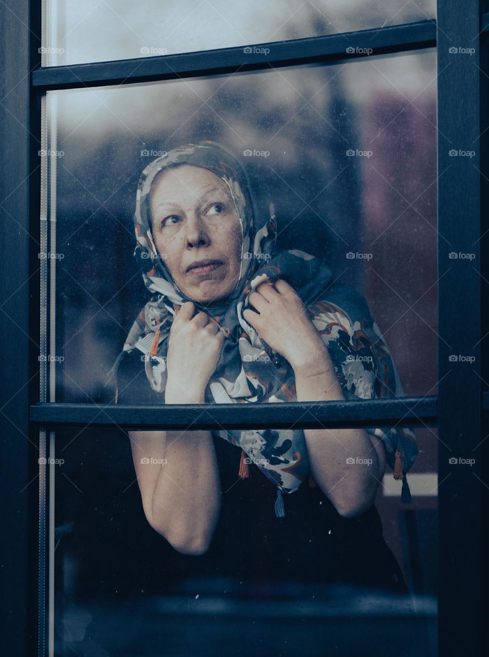 Sad mature woman with scarf on head standing behind dirty window glass