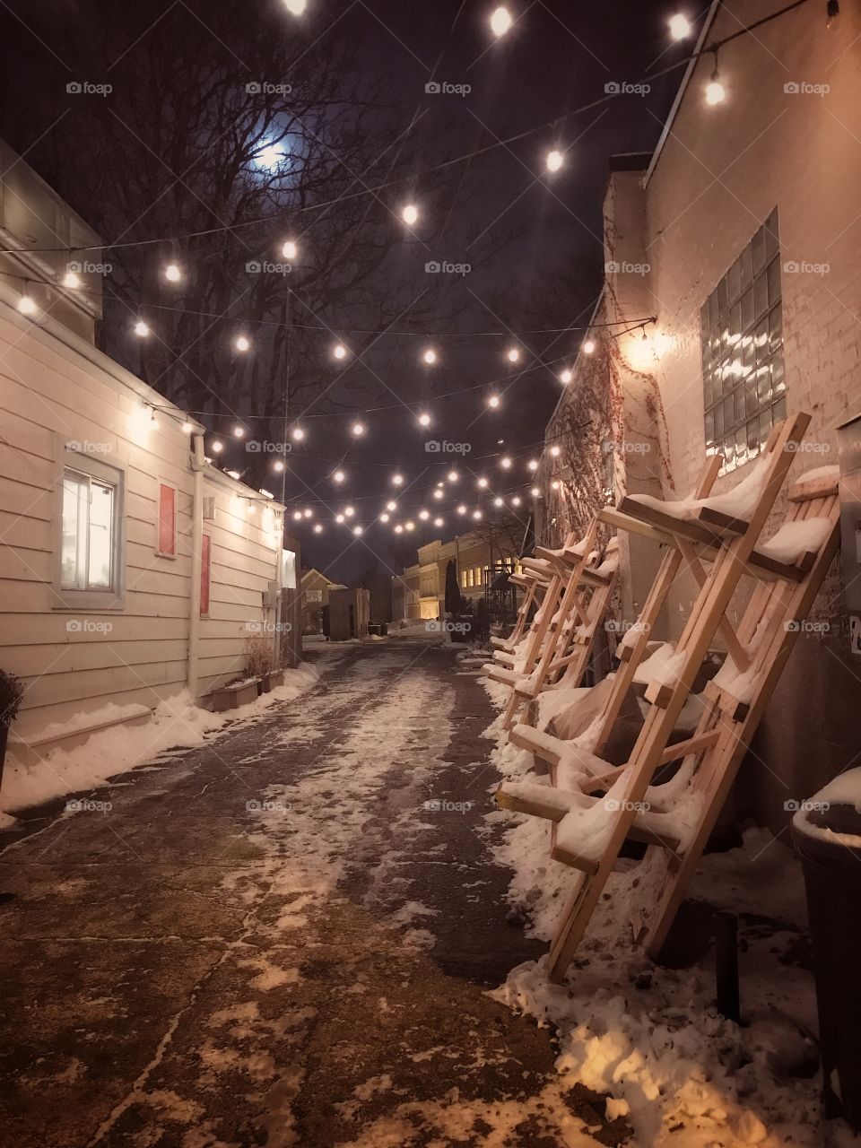 A snowy alley in a small town, bistro lights pair with a glowing moon through the haze of a winter evening in small town America. A line of picnic tables, stacked along the wall since it’s not longer the season for outdoor eating, are draped in snow.
