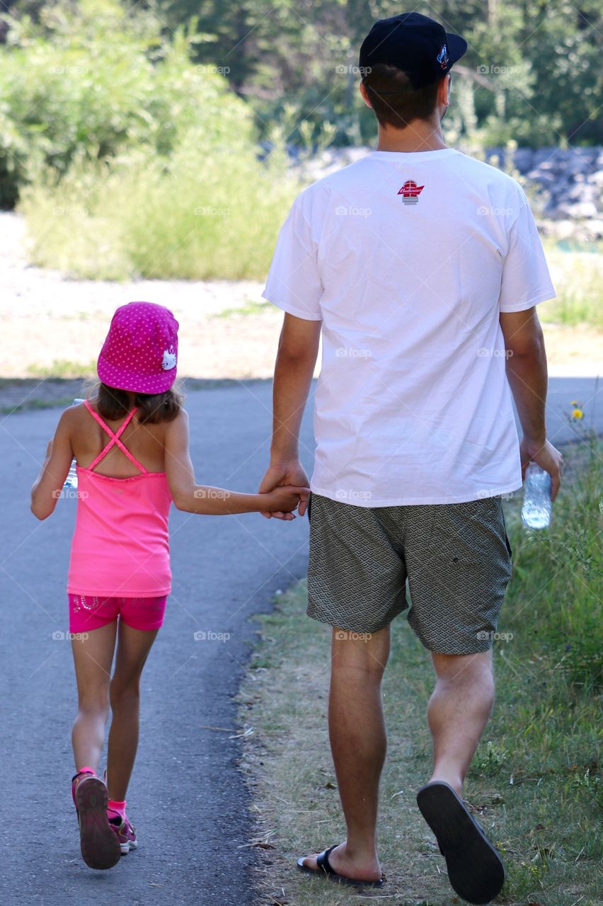 Father holding hands with six year old daughter walking away from camera on path in park on summer day