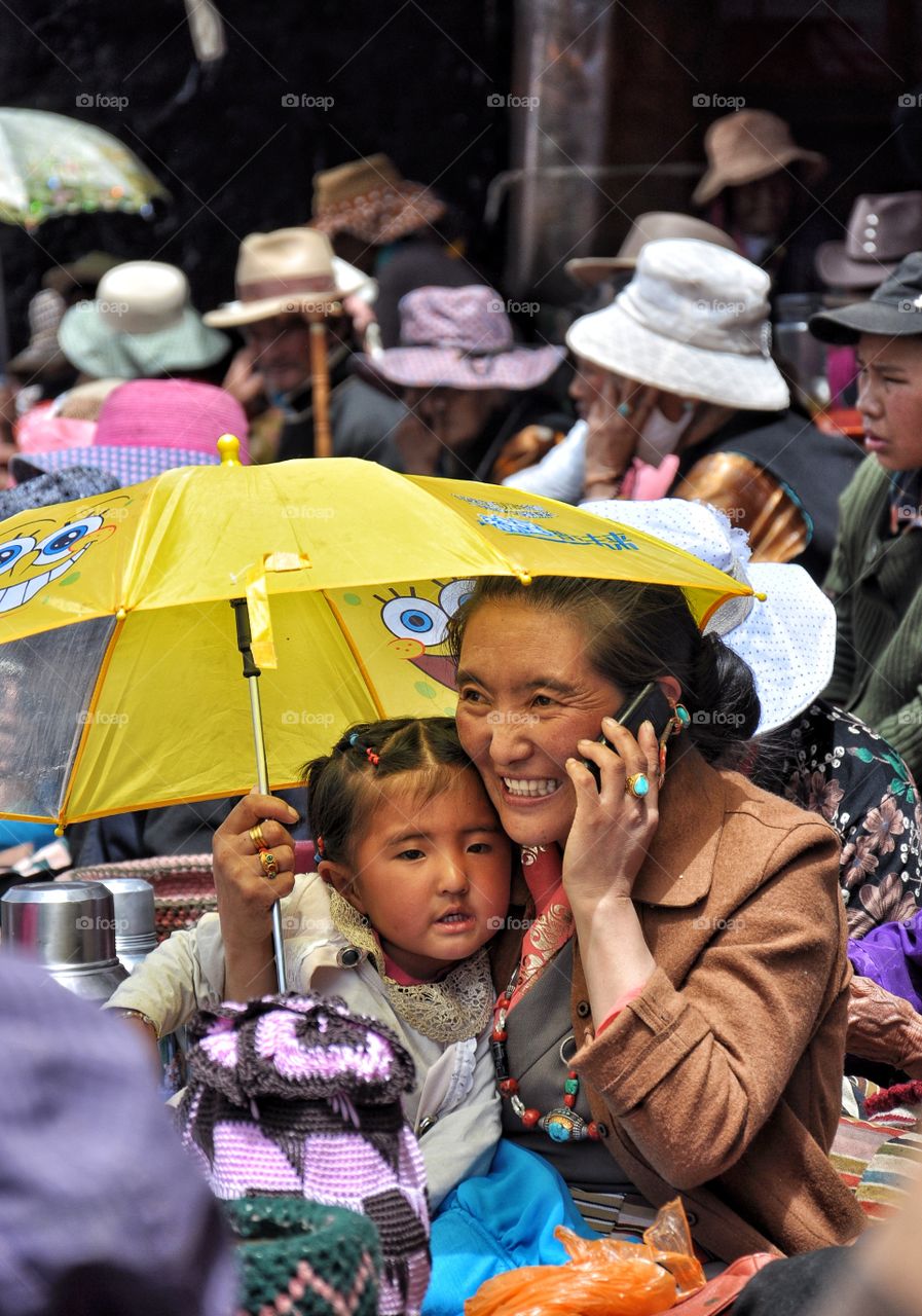 woman speaking on a phone holding girl on her knees under yellow umbrella in the yard of buddhist monastery in tibet