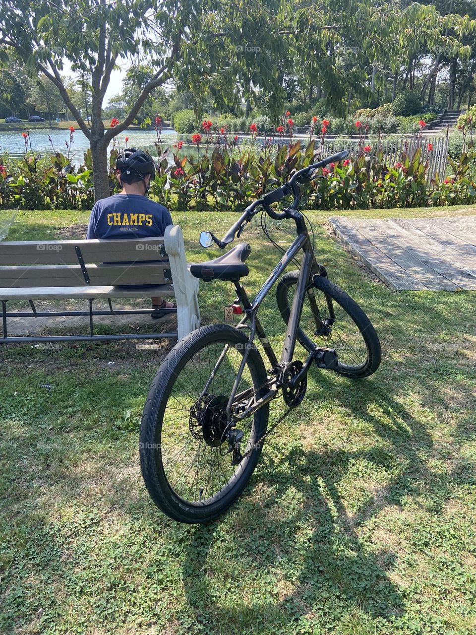 A man sits on a bench next to his bicycle near the lake in Divine Park in Spring Lake, NJ. Flowers and plants surround the lake, and it is a peaceful place to ride or relax.