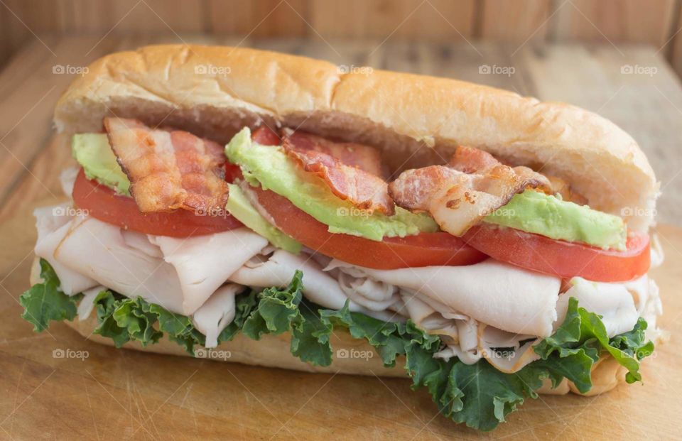 Turkey, bacon, and avocado club submarine sandwich with lettuce and tomatoes served on a wood table with a wood background