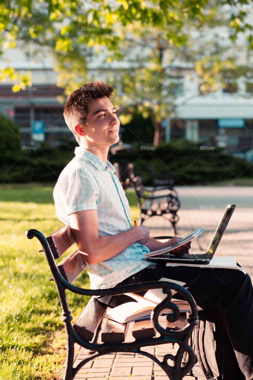 Student working on a laptop using books and notes sitting on a bench in a park. Young boy wearing a blue shirt and dark jeans