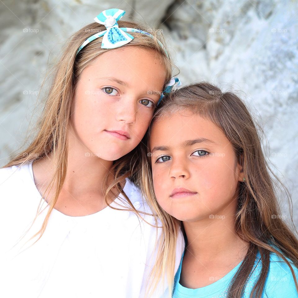 Closeup of two young girls dressed in teal and white standing in repose outside in front of rock cliff formation at beach