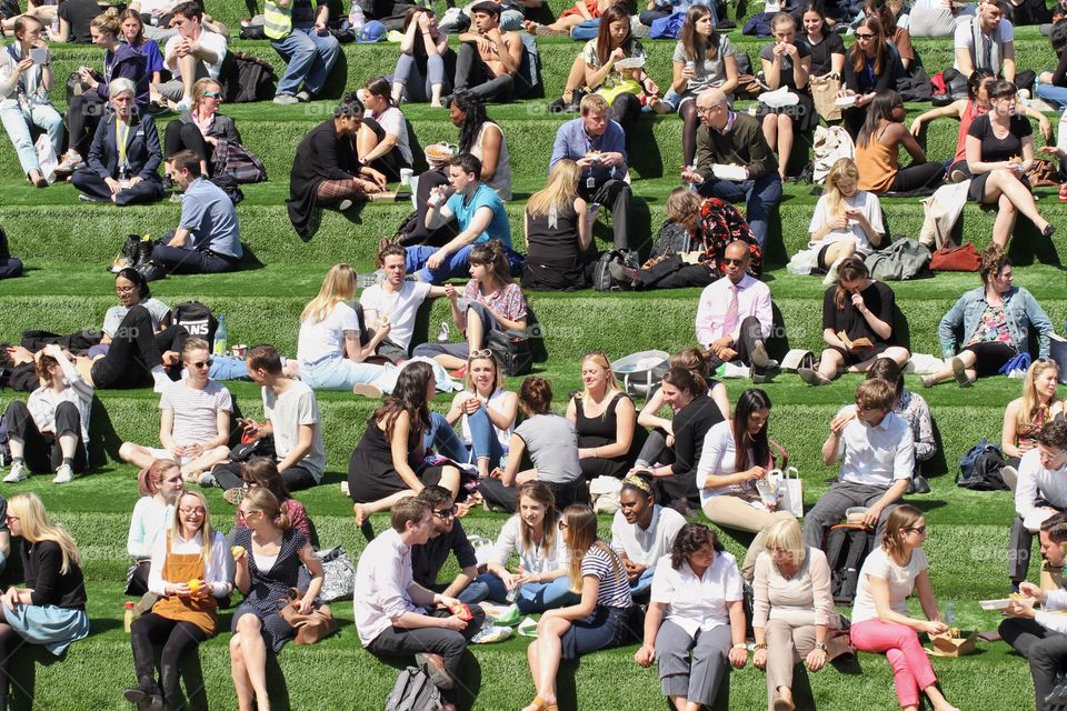 Lots of people enjoying their lunch break in the summer sunshine and relaxing on a grassy hill or bank. 