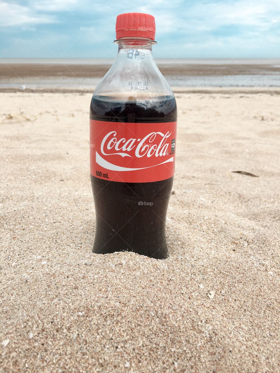 Single bottle of Coca Cola  in the sand set against the backdrop of sky ocean beach 