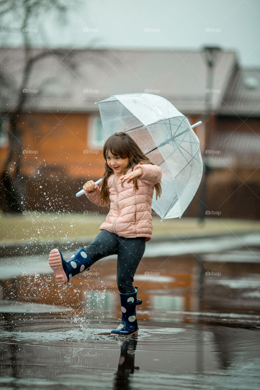 Little girl with umbrella and waterproof boots have fun in puddle 