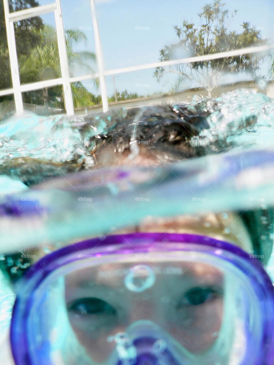 Summer fun! Children swimming in the pool looking at the camera underwater with purple goggles and eyes, seeing above the water splash from jumping in water and enclosure around the pool outside.