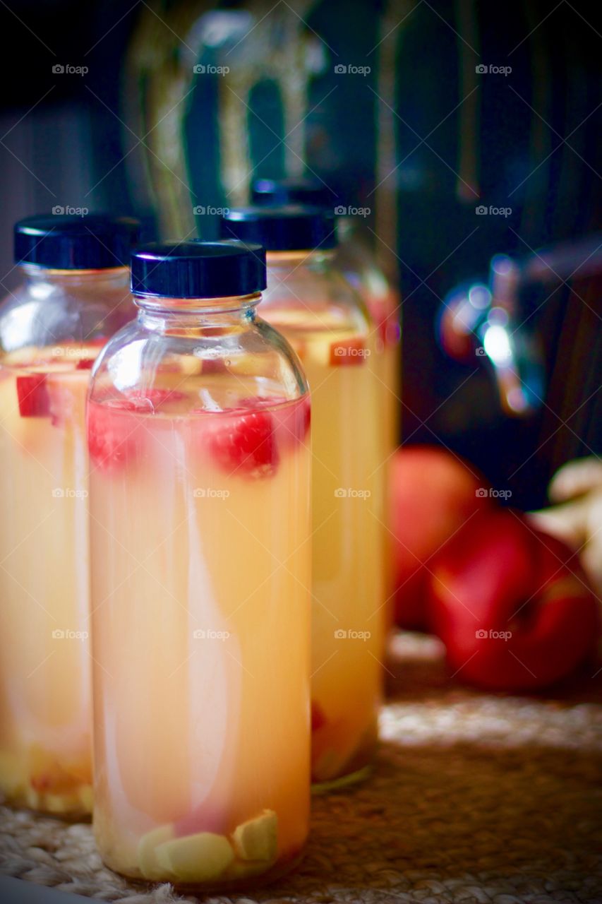 Kombucha, bottled for a second-ferment, flavored with nectarines, raspberries and ginger root slices on a natural fiber mat, whole nectarines and raspberries in a paper carton, stoneware kombucha crock with a stainless steel spigot in the background