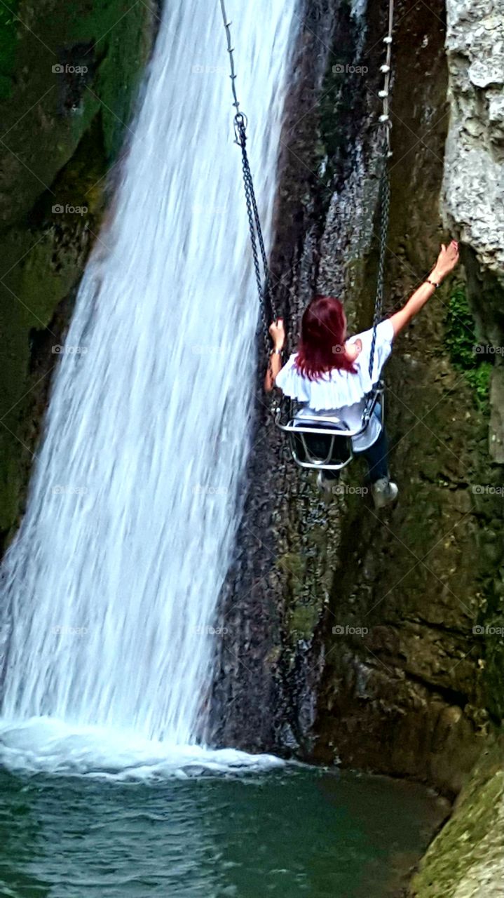 In the cave, young girl near a waterfall flies with the swing to touch the rock wall.