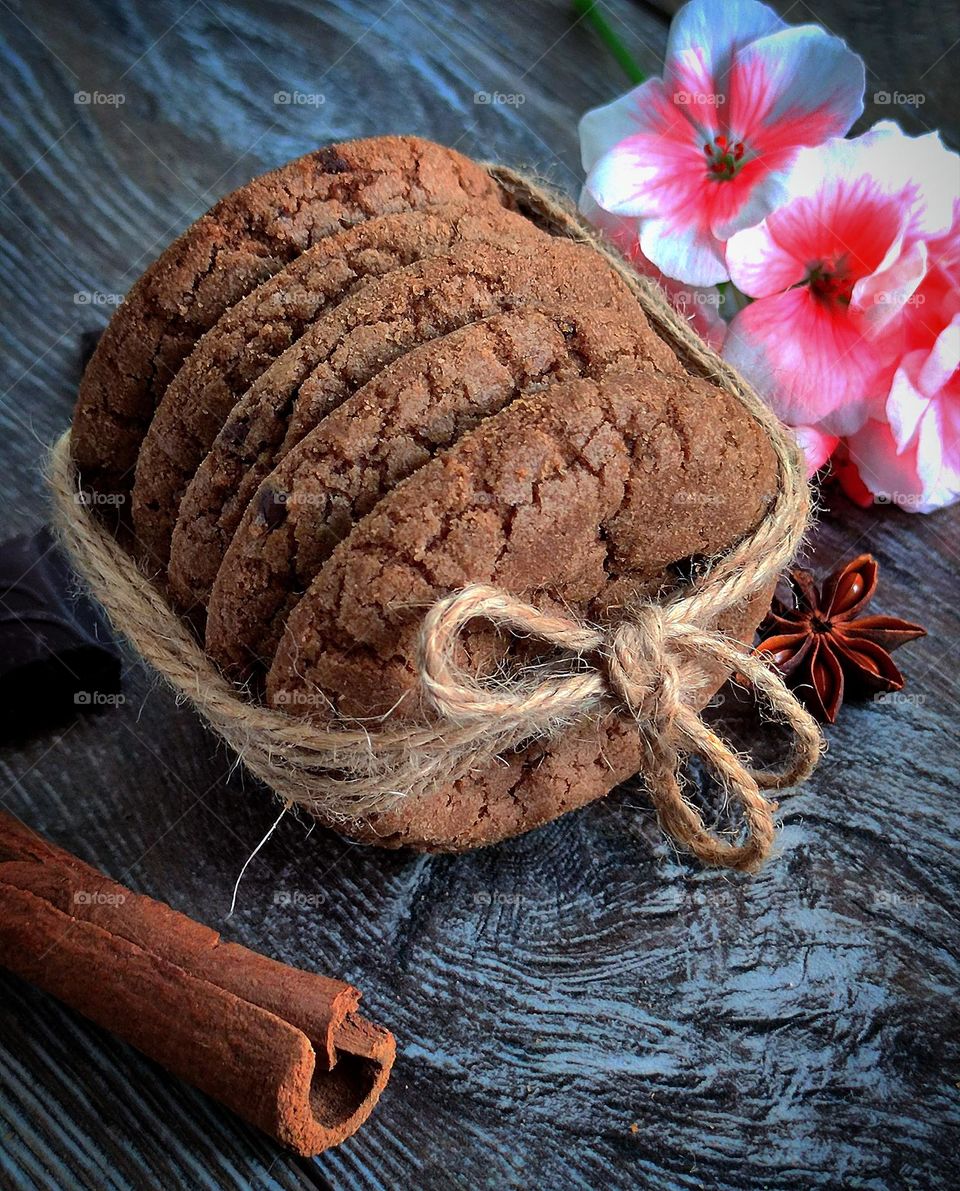 On a wooden background are: a stack of chocolate cookies tied with gray twine, a cinnamon stick, a piece of chocolate, anise flower and pink geranium flowers