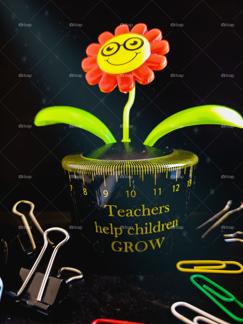 Teacher Appreciation. Still life of a solar plant with magic light shining down. The message  on the solar plant says teachers help children grow and it is surrounded by paper clips and binder clips on a black background.