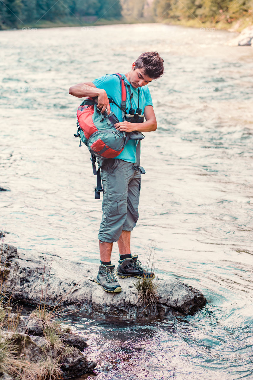 Young boy takes out outdoor mug to take pure water from a river. He is standing on a rock over the river, rests during a hike, spends a vacation on wandering with backpack, he is wearing sport summer clothes