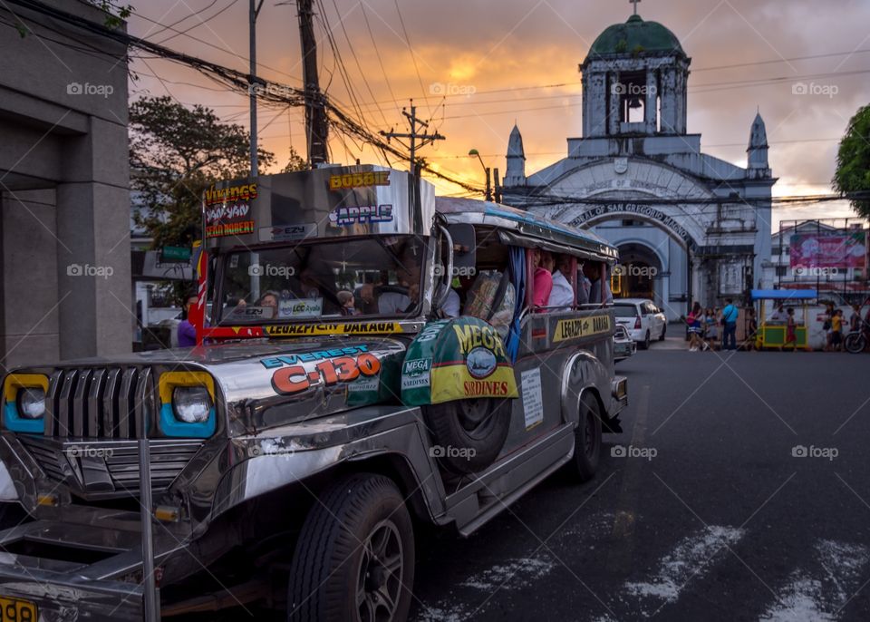 A jeepney, the common mode of public transport in the Philippines, waiting to fill up before setting off in front of a Spanish style Catholic Church at sunset. 