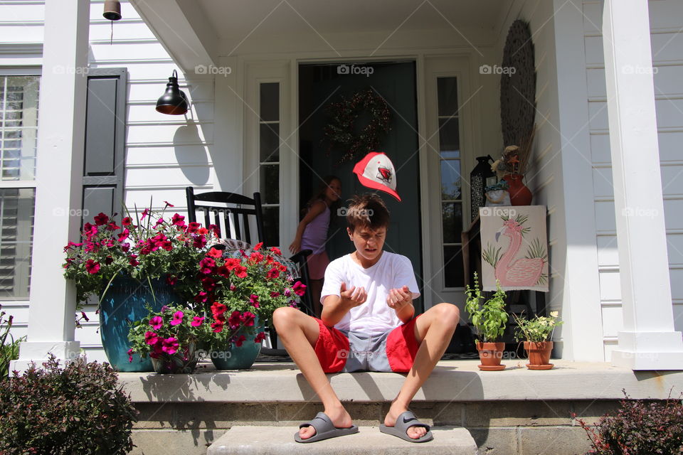 Boy sitting of front stoop while throwing his hat up in air to catch on his head 