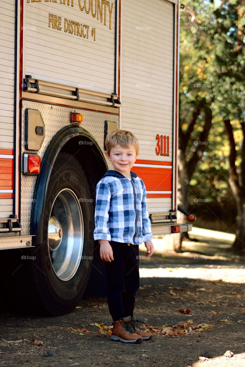 Little boy standing proudly next to a firetruck , fall colors 