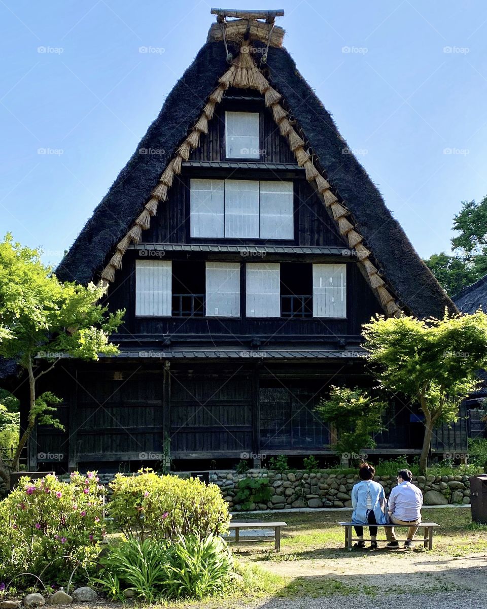 A couple rest on a bench while viewing a Japanese traditional thatched roof house at the Japan Open-Air Folk House Museum.