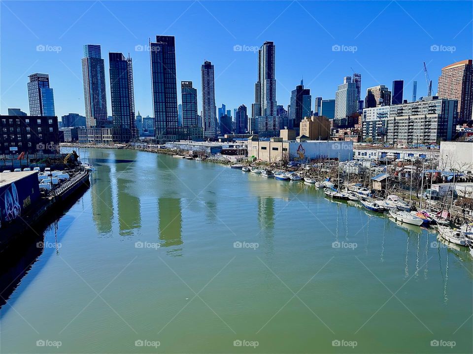 This is „Newtown Creek“ with a great variety of boats seen from the „Pulaski Bridge“ that connects LIC, Queens to „Greenpoint“, Brooklyn. Across the water in the distance behind LIC we see „Manhattan“. 2024. Hypnotic Productions
