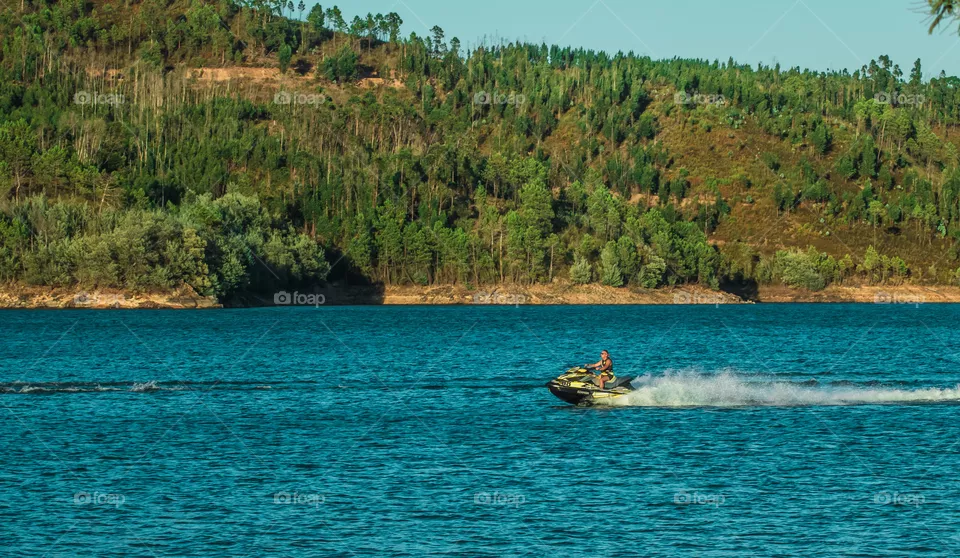 A middle aged man rides a large jet ski along a beautifully blue river, surrounded by green hills, late afternoon on a sunny day