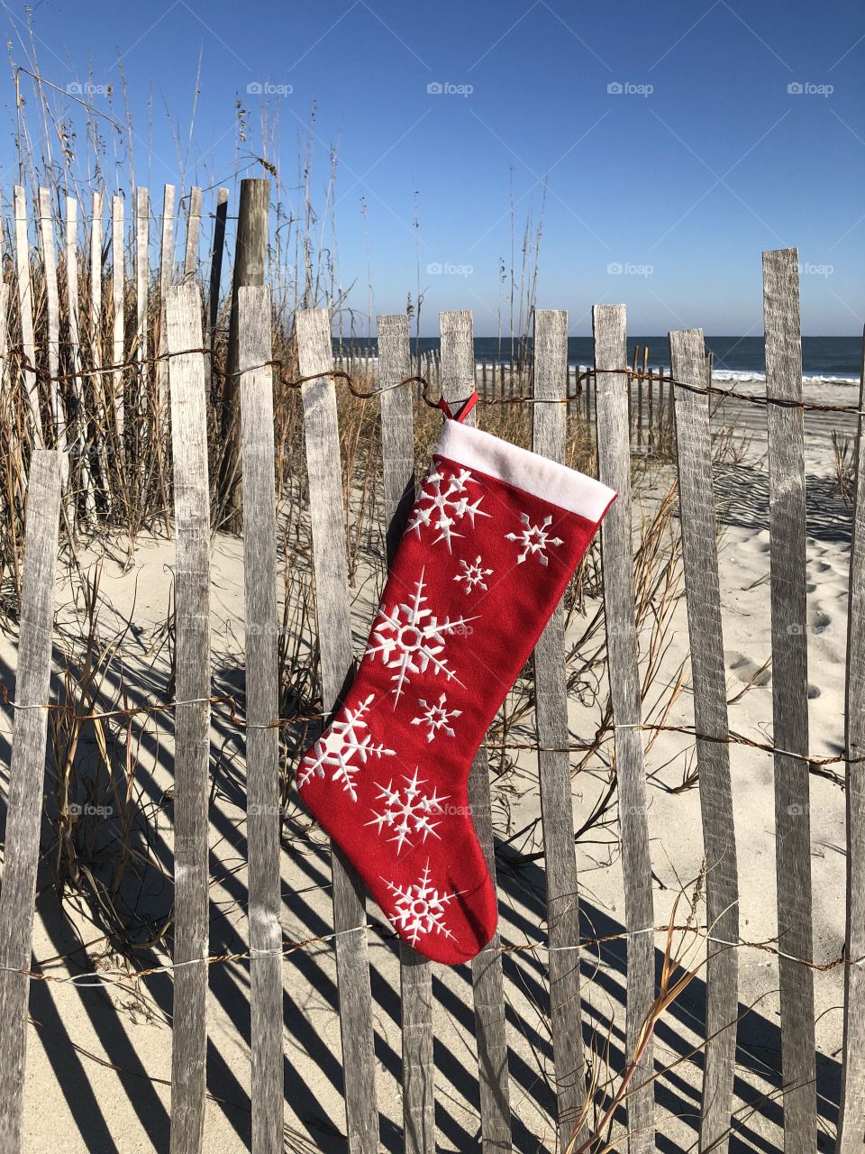 A red Christmas stocking hangs on a beach fence with the Atlantic Ocean in the background. 
