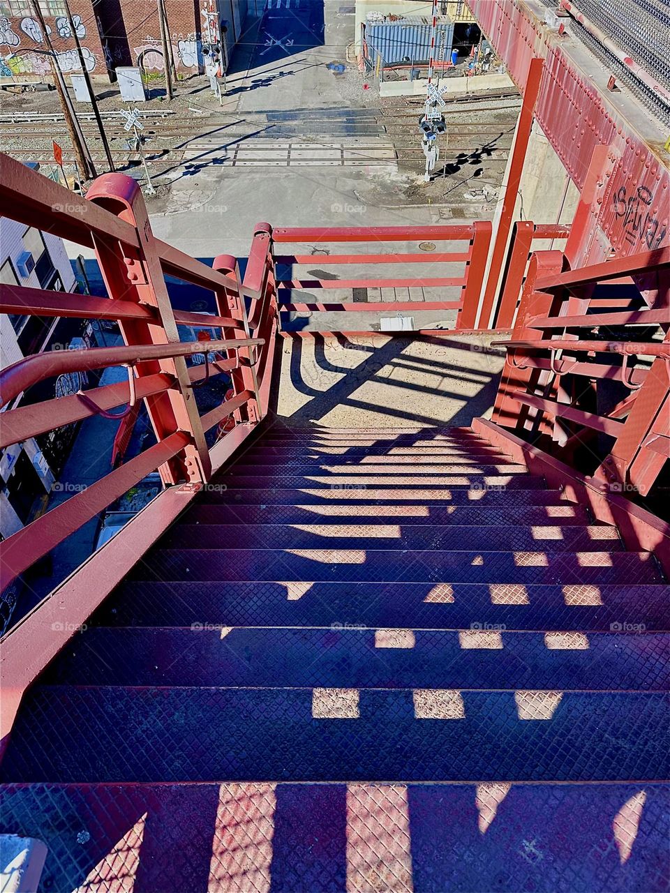 The red metal staircase of the “Pulaski Bridge” at “Newtown Creek” in LIC, Queens has interesting structural design elements. The late afternoon sun often casts dramatic shadows that create intricate interwoven patterns. 2024. Hypnotic Productions