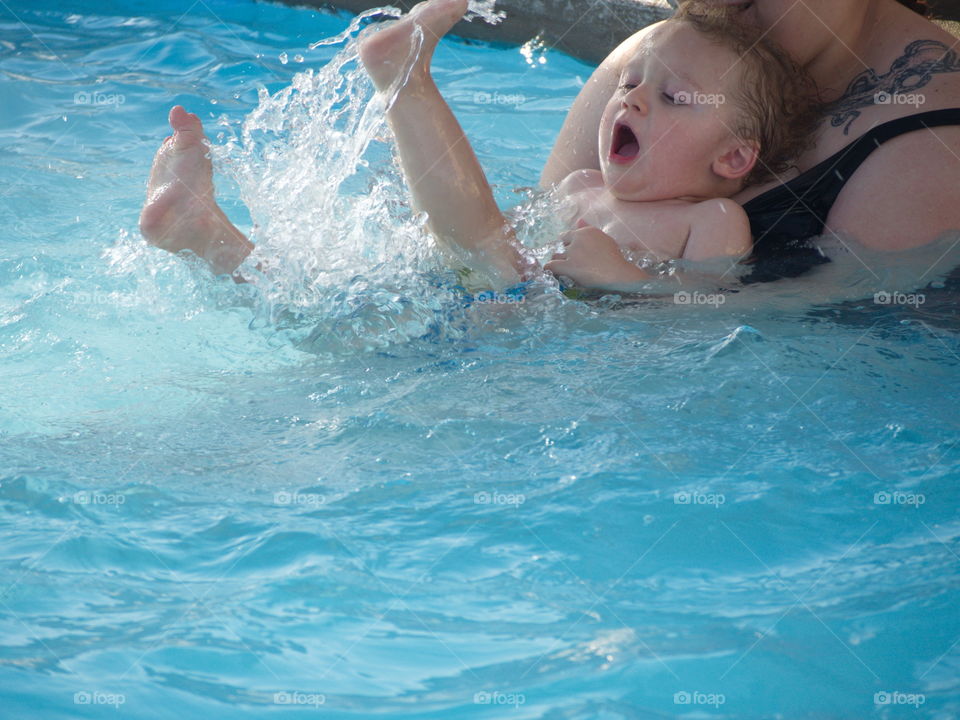 A young toddler boy kicks and splashes in an outdoor pool from the safety of his mothers arms on a sunny summer day. 