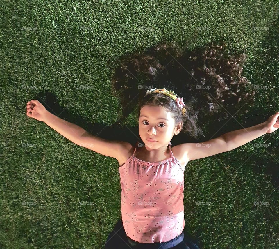 "Embracing the Moment"

A captivating portrait of a young girl lying on lush green grass, her arms wide open as if embracing the world. Her voluminous curly hair fans out like a natural crown, adorned with a delicate floral headband, enhancing her pr