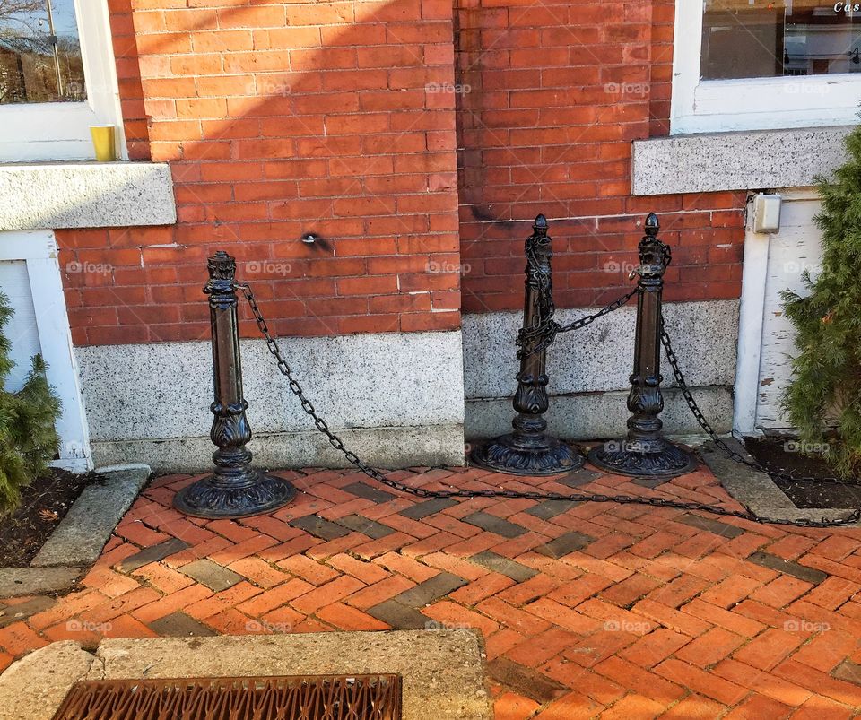 These posts cordon off an eating area outside of a restaurant in an historic district. They are iron and rest on bricks in slanting afternoon light.