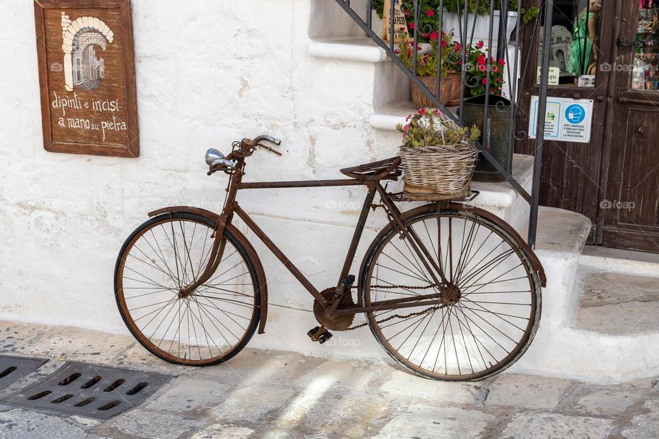 An old and rusty bicycle with a wicker basket and flowers on the back of the trunk stands near a house with a white stone wall and steps leading up on a clear sunny day in the city of Ostuni in Italy, close-up side view.