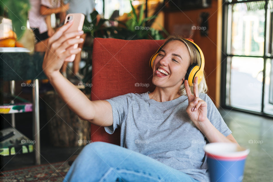 Happy young blonde woman in yellow headphones and grey t-shirt taking selfie in cafe