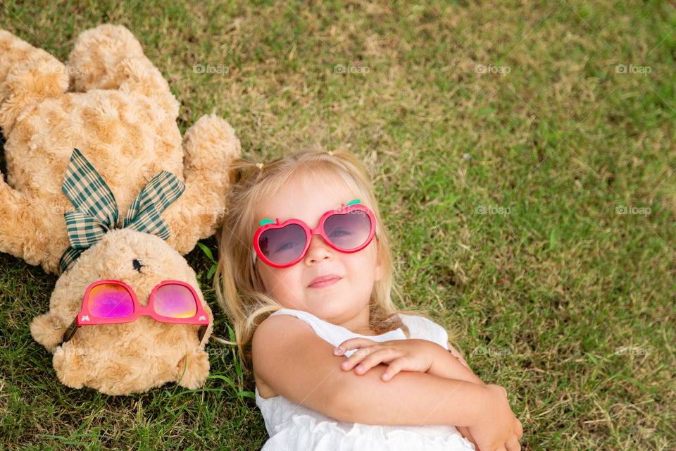 Cute little girl with blonde hair in sunglasses lying on the grass with her teddy bear 