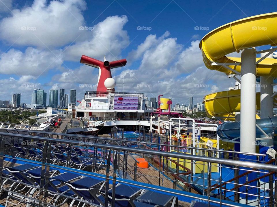 A cruise passenger soaks in all the Carnival Cruise Ship has to offer while waiting to depart from the Miami port. In this picture are blue chairs, a yellow water slide, a kids splash pad, and the infamous Carnival fin surrounded with a blue sky. 