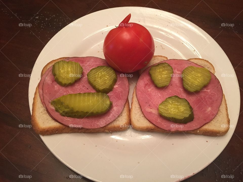 A plate with sandwich bread with meat and pickle faces and a whole garden tomatoe with an arm growing off of it, making this photo rather cute.