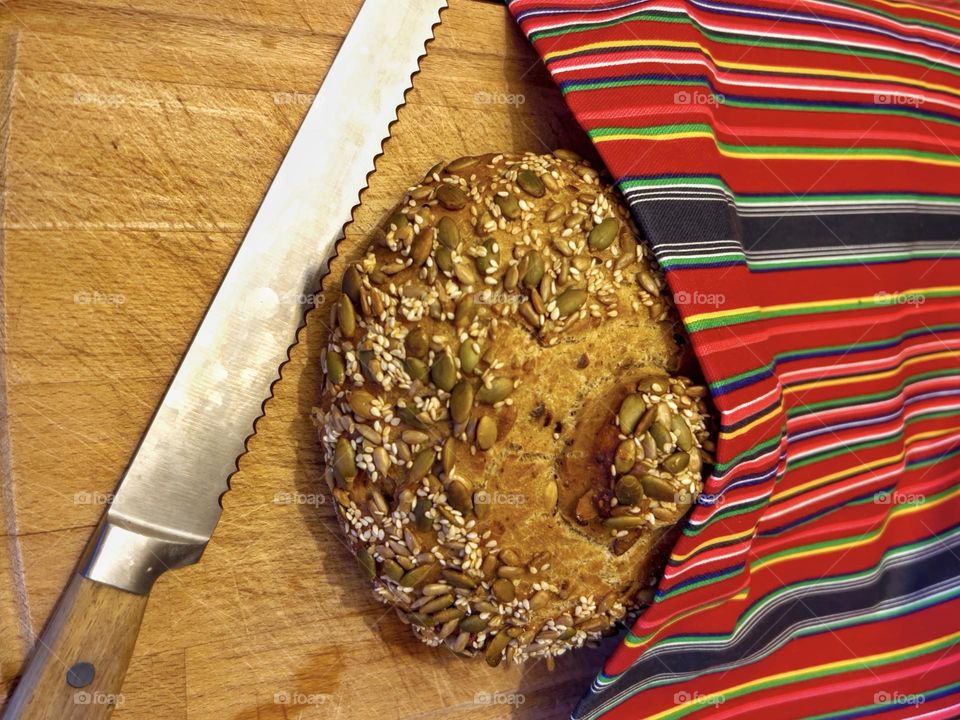 Freshly baked bread lying on a chopping board with a colourful cloth and a bread knife waiting to be cut.