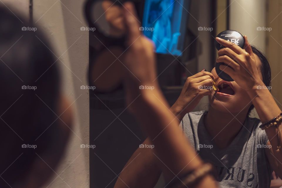 A woman sitting down in front of the mirror while applying her red lipstick, she’s using another small cosmetic mirror to ensure the detail