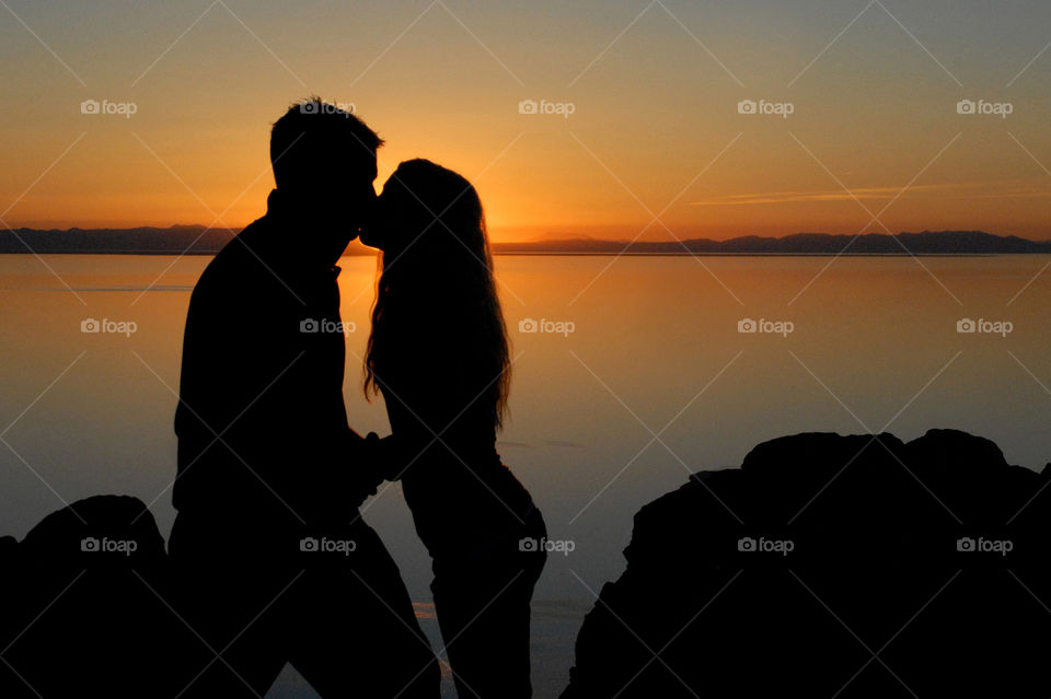 silhouette of a man and woman kissing in the sunset over the horizon of a lake and island in the background - romance