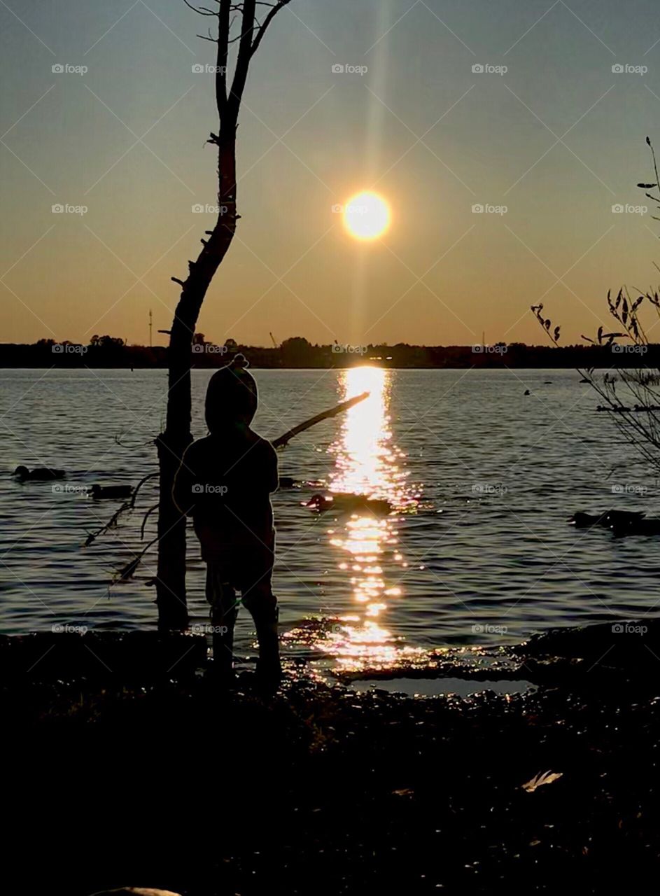 Young boy holding a stick over a lake while the sun sets and ducks swim on the water