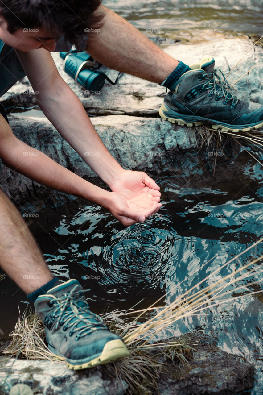 Young boy takes pure water from a river and holds it in the hands. He is sitting on a rock over the river, rests during a hike, spends a vacation on wandering with backpack, he is wearing sport summer clothes