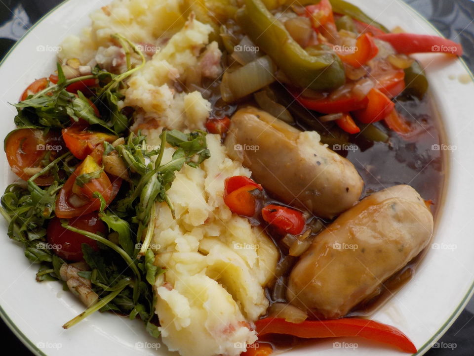Sausages and mash in gravy with peppers and onion, served with tomato and rocket