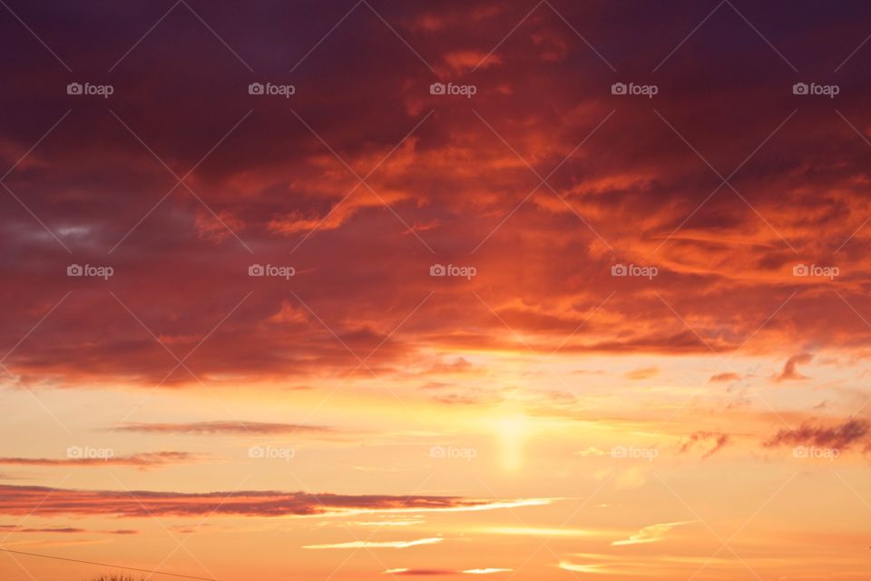 A canopy of brilliantly colored orange and yellow clouds in a pastel-yellow sky with a single vertical ray from the sun